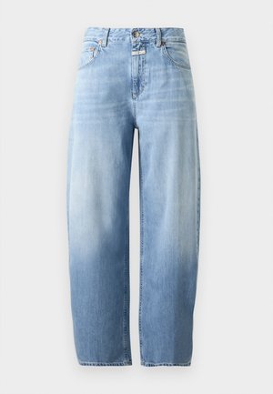 Femme debout portant un blazer beige à carreaux, une chemise grise à col, un jean bleu clair ample et des chaussures plates noires sur fond blanc.