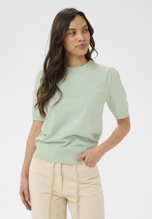 Kobieta o długich ciemnych włosach, ubrana w jasnozielony sweter z krótkim rękawem i beżowe spodnie z fakturą, stojąca z jedną ręką w kieszeni.