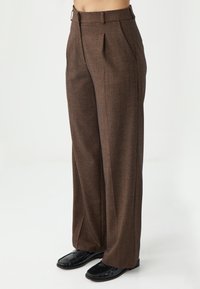 Bruine broek in visgraatmotief met een bredere pijp, voorzien van plooien aan de voorkant, een platte tailleband en een zachte textuur. Gepaard met zwarte schoenen.