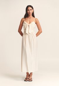 Maxi dress in cream fabric with black polka dots, featuring a bow tie at the front, thin straps, and a flowing silhouette.