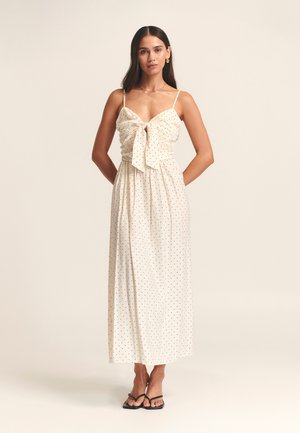 Maxi dress in cream fabric with black polka dots, featuring a bow tie at the front, thin straps, and a flowing silhouette.