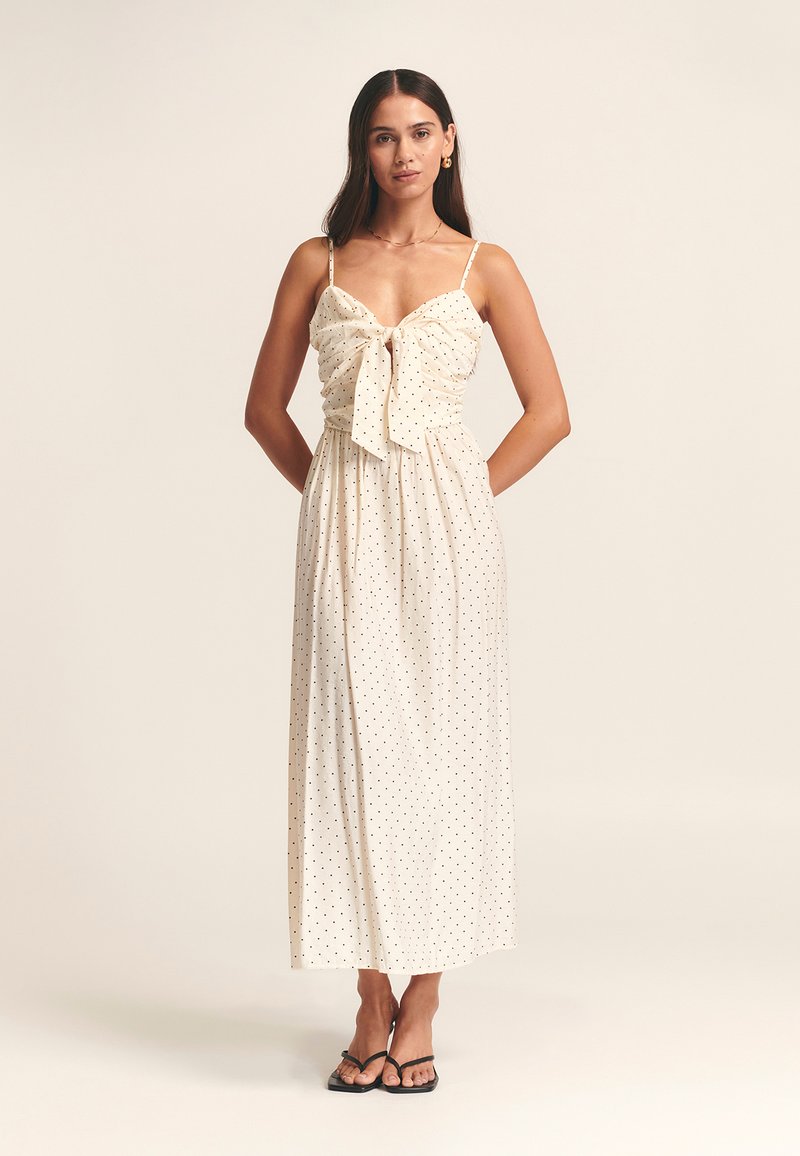 Maxi dress in cream fabric with black polka dots, featuring a bow tie at the front, thin straps, and a flowing silhouette.