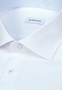 Col de chemise texturé blanc et boutons supérieurs avec étiquette de marque "seidensticker" visible à l'intérieur du col.