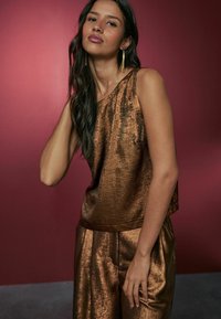 Bronze metallic sleeveless top with a loose fit, paired with matching loose trousers, set against a pink background.