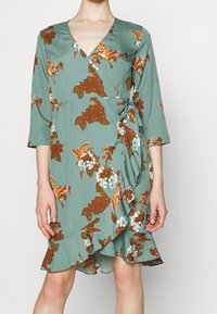 V-aukko, kolme neljännesosaa hihoja ja röyhelöhelma, vihreä kangas, jossa ruskeita ja valkoisia kukkakuoseja.