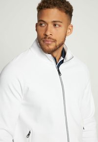 Chaqueta blanca con cremallera y cuello alto, con detalles en negro. Presenta pequeños bolsillos laterales con cremallera y un tejido texturizado.