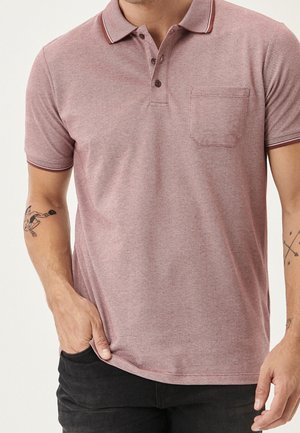 Uomo che indossa una polo a maniche corte di colore rosso chiaro con taschino e due tatuaggi visibili sulle braccia, mano nella tasca dei pantaloni neri.