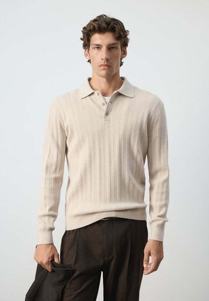 Jeune homme aux cheveux bouclés portant un pull polo en maille beige et un pantalon foncé, tenant une veste noire dans la main gauche devant un fond uni.