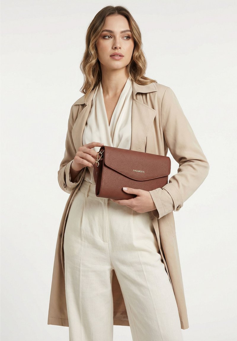Mujer con abrigo beige y conjunto blanco sosteniendo un bolso de mano marrón de cuero Lazarotti sobre un fondo liso.