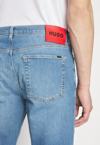 Vaqueros de denim azul claro con un parche de cuero rojo etiquetado "HUGO" en la cinturilla, que cuentan con bolsillos traseros y sutiles detalles de costura.