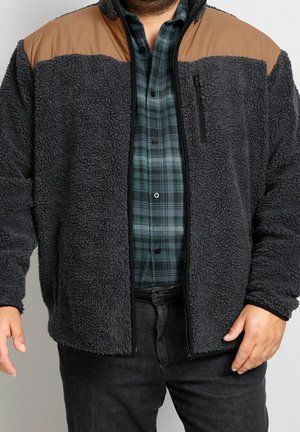 Uomo che indossa una giacca in pile sherpa nera con pannelli beige sulle spalle, sopra una camicia a quadri verde e nera e pantaloni neri.