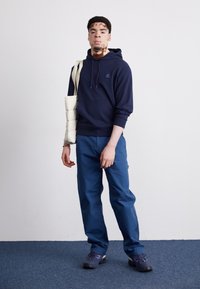 Marineblaue Kapuzenjacke mit Logo, kombiniert mit blauen Hosen. Die Accessoires bestehen aus einer beigefarbenen Schultertasche und dunklen Sneakers. Einfaches, lässiges Design.