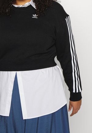 Osoba nosząca czarną bluzę Adidas z białymi paskami na rękawach, pod którą ma białą zapinaną na guziki koszulę i niebieską spódnicę, stoi na białym tle.