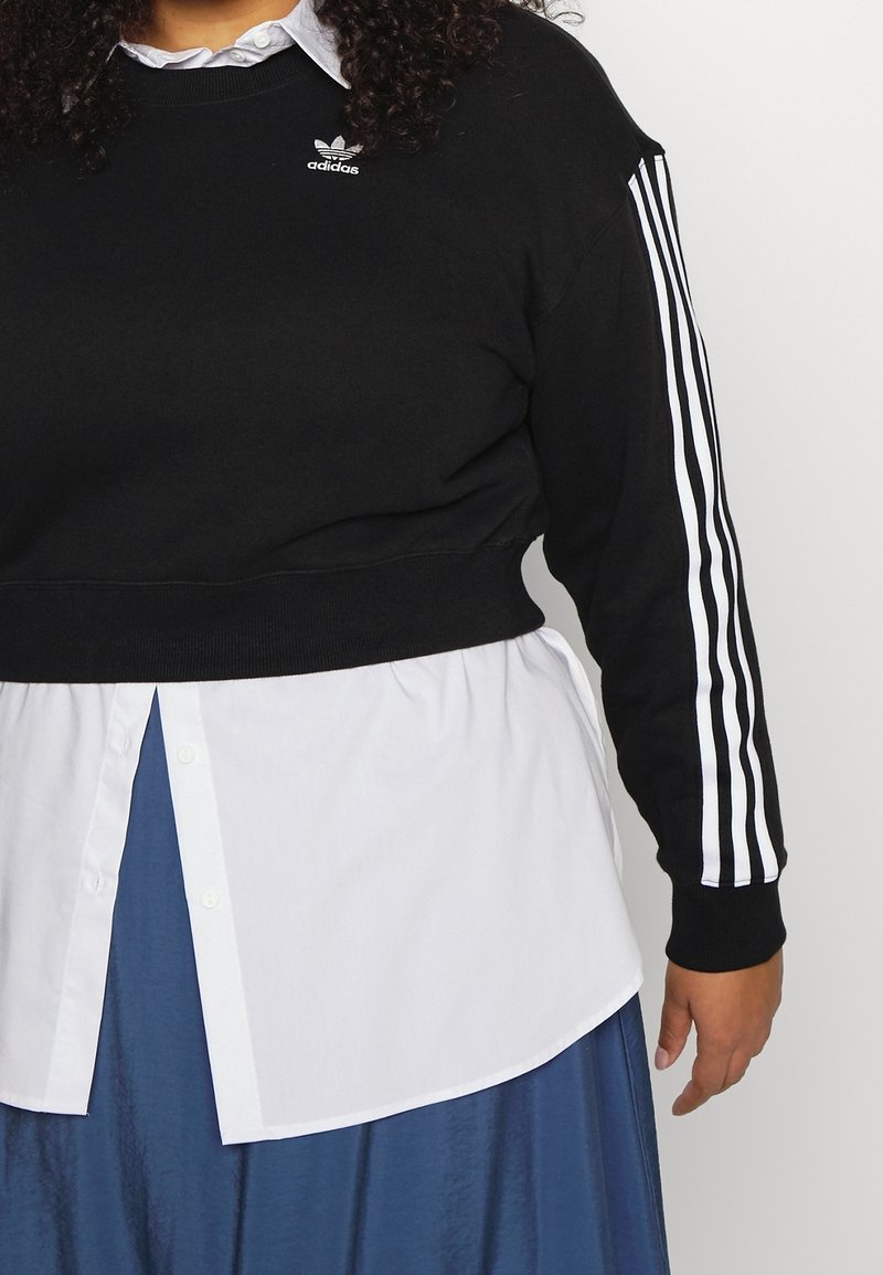 Personne portant un sweat Adidas noir avec des bandes blanches sur les manches, par-dessus une chemise blanche boutonnée et une jupe bleue, se tient devant un fond blanc.