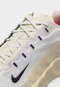 Gros plan sur une basket Nike blanche et crème avec une couture ondulée sur le côté, le logo Swoosh noir et des lacets blancs sur une tige perforée.