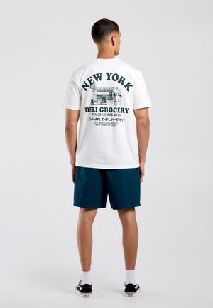 Man met zijn rug naar de camera, draagt een wit T-shirt met een groene New Yorkse delicatessenwinkel-afbeelding en marineblauwe korte broek voor een effen achtergrond.