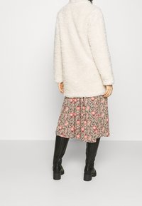 Hvid, fluffet frakke med høj krave, båret over en blomsterprintet kjole i pink og grøn, kombineret med knælange sorte støvler.