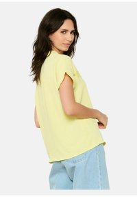 Top corto giallo chiaro, leggero e dall'aspetto sciolto, con una finitura texturizzata e un dettaglio in pizzo. Abbinato a jeans azzurri chiari a vestibilità rilassata.