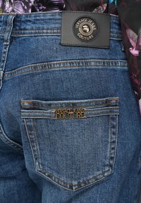 Plave traperice imaju crnu kožnu oznaku na struku s okruglim metalnim kopčanjem. Stražnji džep uključuje zlatnu oznaku "VERSACE JEANS COUTURE".