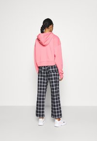 Kobieta stojąca tyłem, ubrana w różową bluzę z kapturem, czarno-białe kraciaste spodnie, białe trampki oraz z warkoczami.