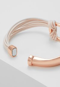Armband i roséguld med flera sammanflätade vita snören, en rektangulär detalj med en ljus infälld del och en slät, polerad yta.