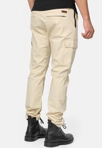 Pantaloni cargo beige con tasche laterali, tessuto testurizzato, vestibilità su misura e stivaletti neri con lacci, visti da dietro.
