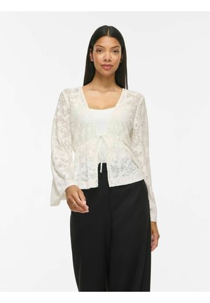 Kvinde med langt mørkt hår iført en hvid blonde cardigan over en hvid top og løse sorte bukser, stående mod en ensfarvet baggrund.