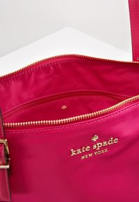 Jasnoróżowa torebka Kate Spade New York z złotym zamkiem błyskawicznym i logo, z otwartą główną komorą i wewnętrzną kieszenią na zamek.