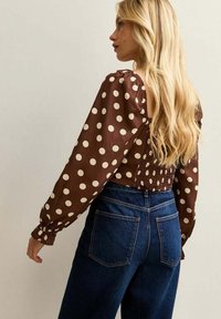 Brun blus med prickar och puffärmar, smockad bak, kombinerad med högmidjade mörka jeans. Texturen ser mjuk ut, med elastiska manschetter.