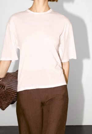 Person wearing a plain white short-sleeve t-shirt and brown pants, holding a textured brown woven clutch with their left hand.
