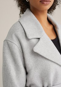 Palton gri deschis din amestec de lână, cu guler decupat, mâneci lungi și un design structurat. Textură moale cu detalii vizibile de cusături.