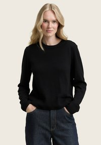 Pull en maille noir à col rond et manches longues ; présente des bords-côtes aux poignets et à l'ourlet ; porté avec un jean bleu. Design simple et classique.