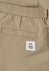 Bežīgas bikses ar elastīgu jostu, ar aizmugurējo kabatu un baltu birku ar melnu uzrakstu "M 73". Gluda, teksturēta auduma.