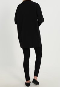 Černá oversized bunda s hladkou texturou, kulatým výstřihem a sníženými rameny, kombinovaná s upnutými černými kalhotami a plochými botami.