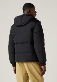 Black puffer jacket with a hood, matte texture, structured seams, elastic cuffs, and a cropped length, paired with beige corduroy pants.