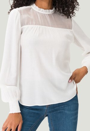 Kobieta ubrana w białą bluzkę z długim rękawem z przezroczystym karczkiem, marszczonym dekoltem i marszczonymi mankietami, w połączeniu z niebieskimi dżinsami, stoi z ręką na biodrze.