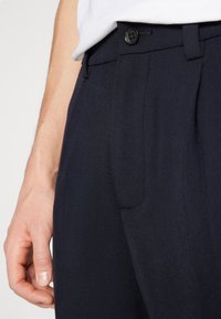 Pantalons navy en tissu lisse, dotés d'une fermeture à bouton, de poches latérales et de détails de pli net. Vue rapprochée de la taille et de la poche.