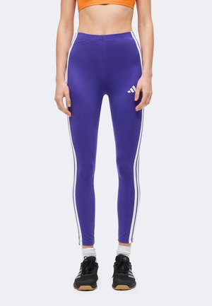 Γυναίκα με μωβ κολάν Adidas με λευκές ρίγες, μαύρα αθλητικά παπούτσια, λευκές κάλτσες και πορτοκαλί τοπ, στέκεται μπροστά σε λευκό φόντο.