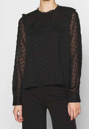 Mujer lleva una blusa negra translúcida de manga larga con textura de lunares y ribete de encaje, combinada con pantalones negros, de pie contra un fondo liso.