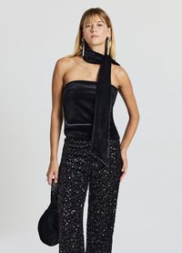 Schwarzes trägerloses Top aus samtartigem Material mit langer Schleife, kombiniert mit schwarzen, mit Pailletten verzierten Hose mit weitem Bein. Accessoire: kleine schwarze Tasche.