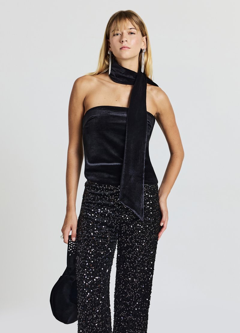 Schwarzes trägerloses Top aus samtartigem Material mit langer Schleife, kombiniert mit schwarzen, mit Pailletten verzierten Hose mit weitem Bein. Accessoire: kleine schwarze Tasche.