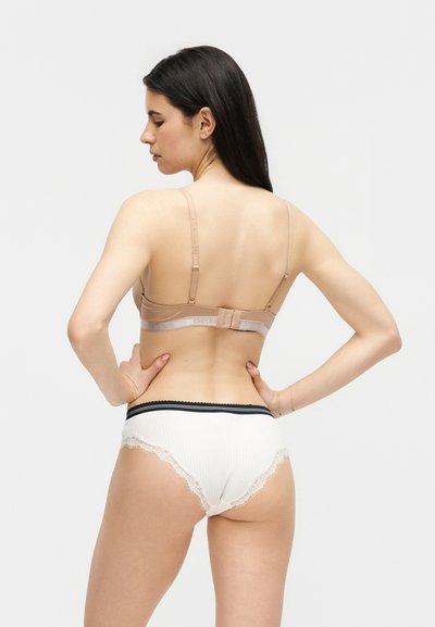 Kvinne med langt mørkt hår iført en beige BH og hvit truse med blonder, stående med hendene på hoftene, vendt bort fra kamera mot en ensfarget bakgrunn.