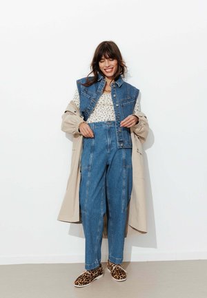 Femme souriante, portant un gilet et un pantalon en denim, un haut à fleurs, un trench beige et des baskets imprimé léopard, debout contre un mur blanc.