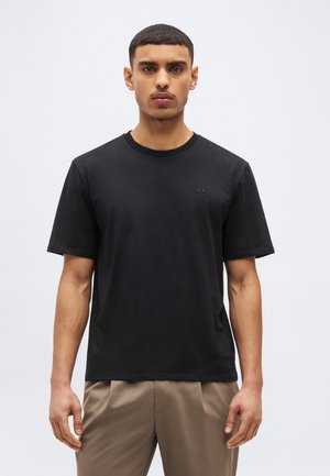 Νεαρός άνδρας με κοντά σκούρα μαλλιά, φοράει απλό μαύρο μπλουζάκι με στρογγυλή λαιμόκοψη και μπεζ παντελόνι, στέκεται μπροστά σε ανοιχτό γκρι φόντο.