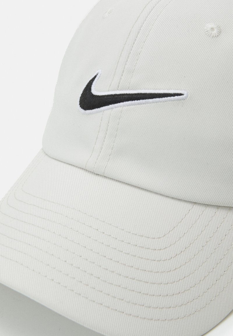 Gorra de béisbol blanca hecha de algodón, con el logotipo bordado de Nike en negro en la parte frontal. Incluye detalles de costura visibles.