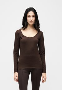 Γυναίκα με μακριά σκούρα μαλλιά, φορώντας εφαρμοστό καφέ μπλουζάκι με μακριά μανίκια και καφέ παντελόνι, στέκεται μπροστά σε λευκό απλό φόντο.