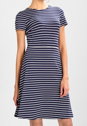 Frau trägt ein marineblaues Kleid mit weißen horizontalen Streifen, kurzen Ärmeln und taillierter Taille und steht vor einem einfarbigen Hintergrund.