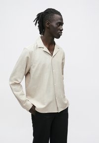 Beżowa koszula z krótkim rękawem o subtelnym wzorze, z pojedynczą kieszenią na piersi i wykładanym kołnierzem, zestawiona z czarnymi spodniami. Gładka faktura.