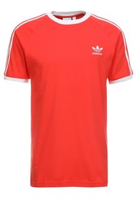 Punainen puuvillainen t-paita, jossa on valkoinen kaulus ja hihansuut, valkoiset kolmiraidat olkapäillä ja Adidas-logo rinnassa.