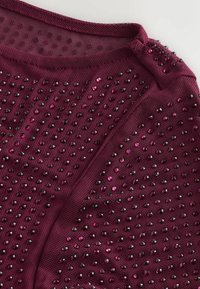Burgunderfarbener Stoff mit einer strukturierten Oberfläche, der mit verstreuten schwarzen und rosa Perlen verziert ist. Der Ausschnitt ist glatt und hat ein kleines Verschlussdetail.
